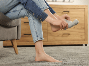Person in jeans sits barefoot on a carpet, about to put on a gray sock in a living room setting.